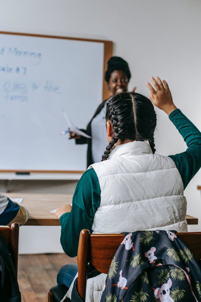 Smiling African American female teacher standing near whiteboard and looking at schoolgirl raising hand